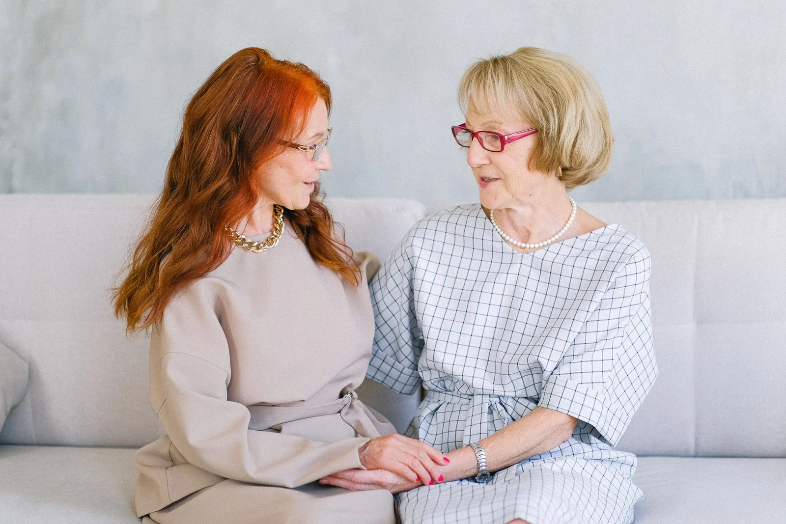 Two older women holding hands and talking on a sofa.