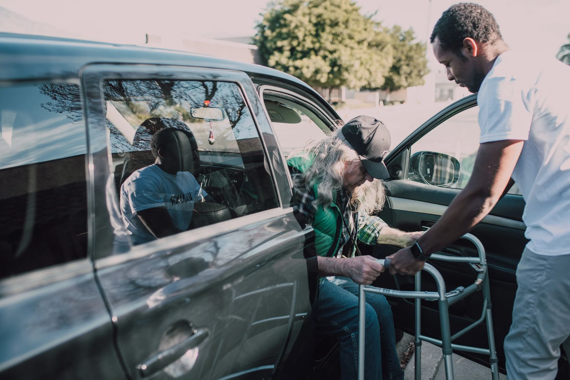 An older adult using a walker receiving assistance beside a vehicle.