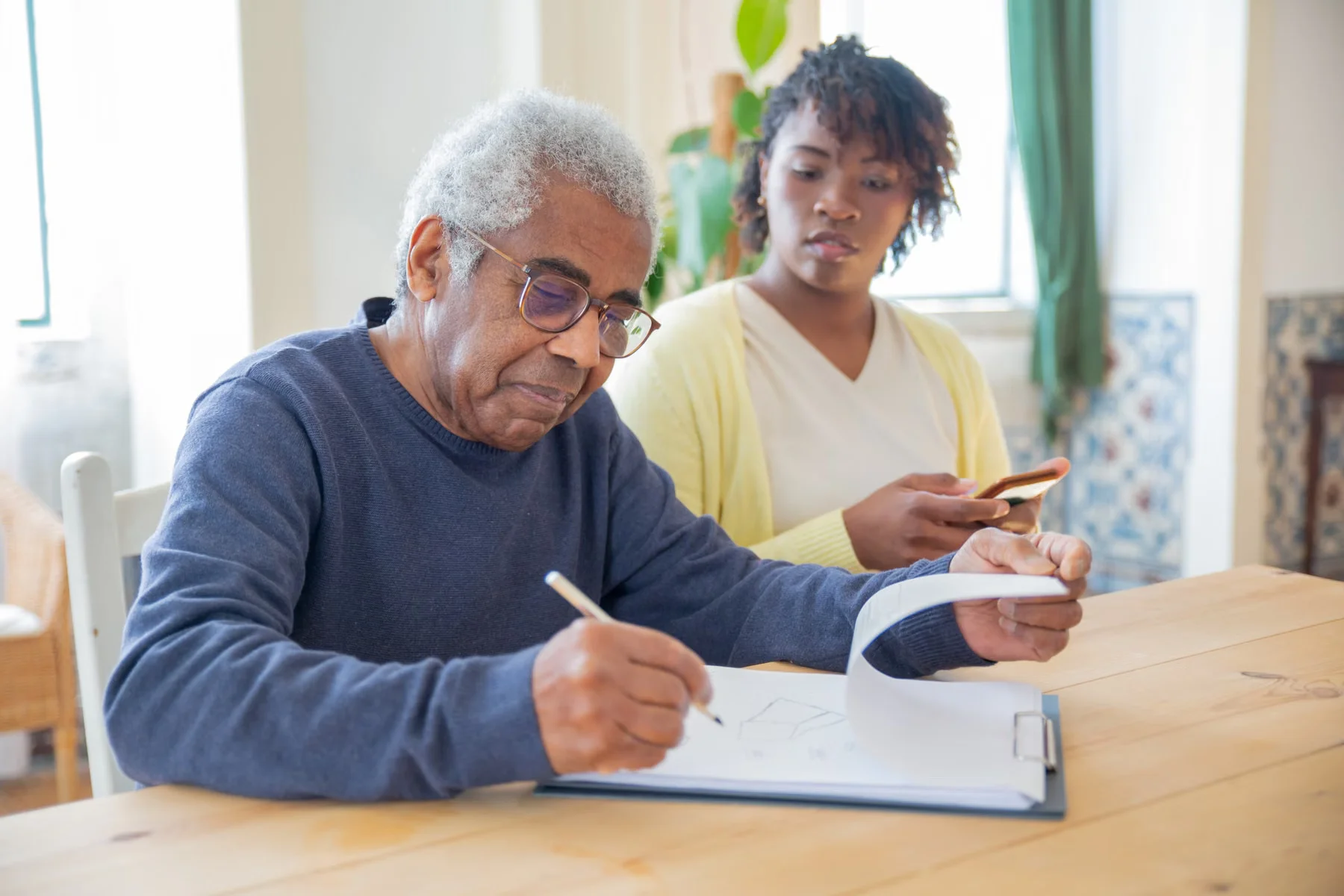 An older man writing in a notebook with a caregiver nearby.
